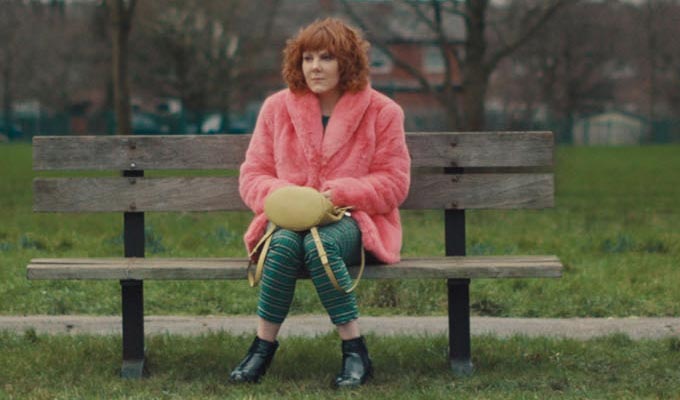 Sophie as Alma, sitting on a park bench in a vivid pink fur coat