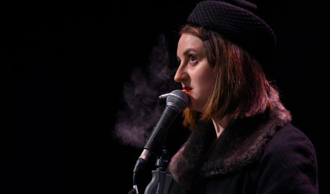 Lorna Rose on stage, in character in long trenchcoat and smoking a cigarette